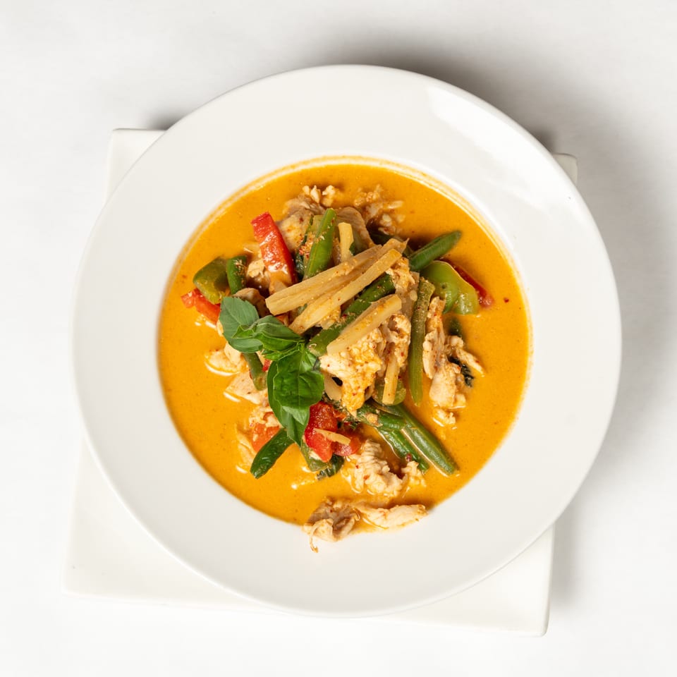 Overhead view of orange curry with chicken, green beans, bamboo shoots, peppers, and basil in a white bowl.