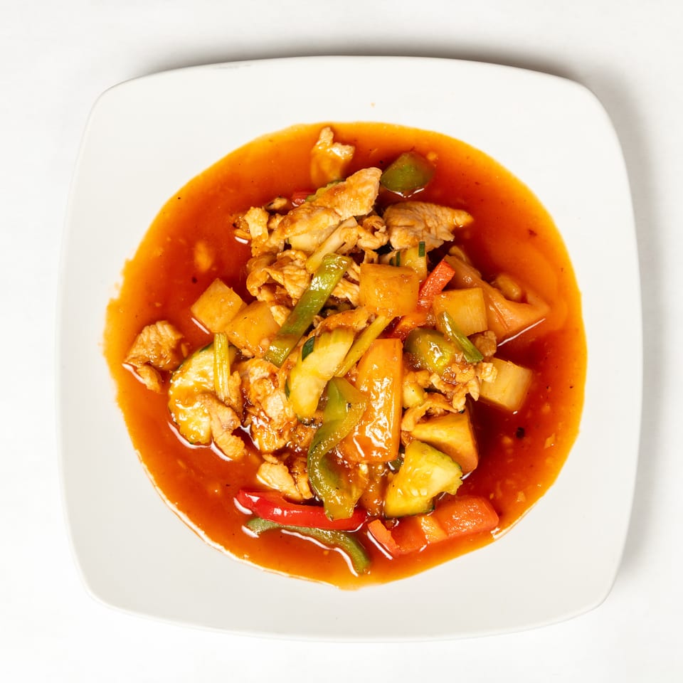 Stir-fried chicken with pineapple, bell peppers, carrots, and vegetables in a glossy red sauce on a white plate.