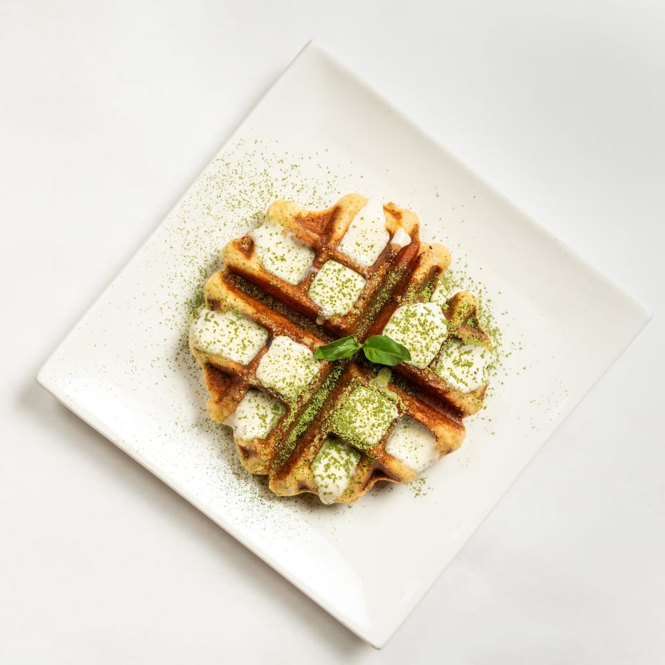 Golden waffles topped with cream, green matcha powder, and fresh basil on a white square plate.