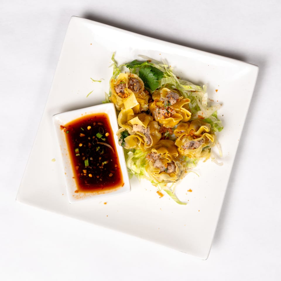 Steamed pork dumplings garnished with chili flakes and herbs on shredded lettuce, served with spicy dipping sauce.