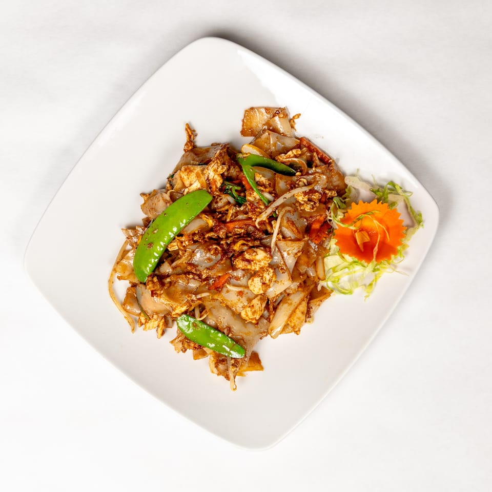 Stir-fried wide rice noodles with chicken, vegetables, and eggs, served with lettuce and carved carrot garnish.